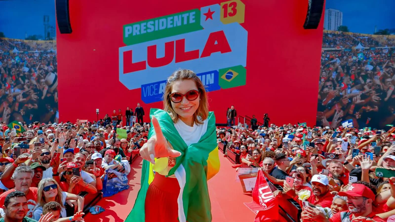 Janja com bandeira do Brasil em palco de campanha com painel Presidente Lula, foto de arquivo do lançamento da campanha em agosto