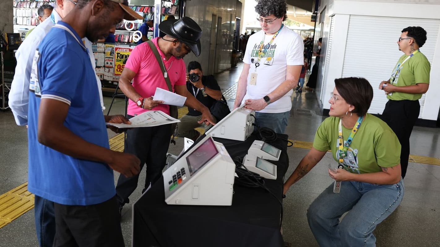 Servidores do TSE, de colete verde, demonstram para eleitores diversos como votar em uma urna eletrônica na Rodoviária de Brasília