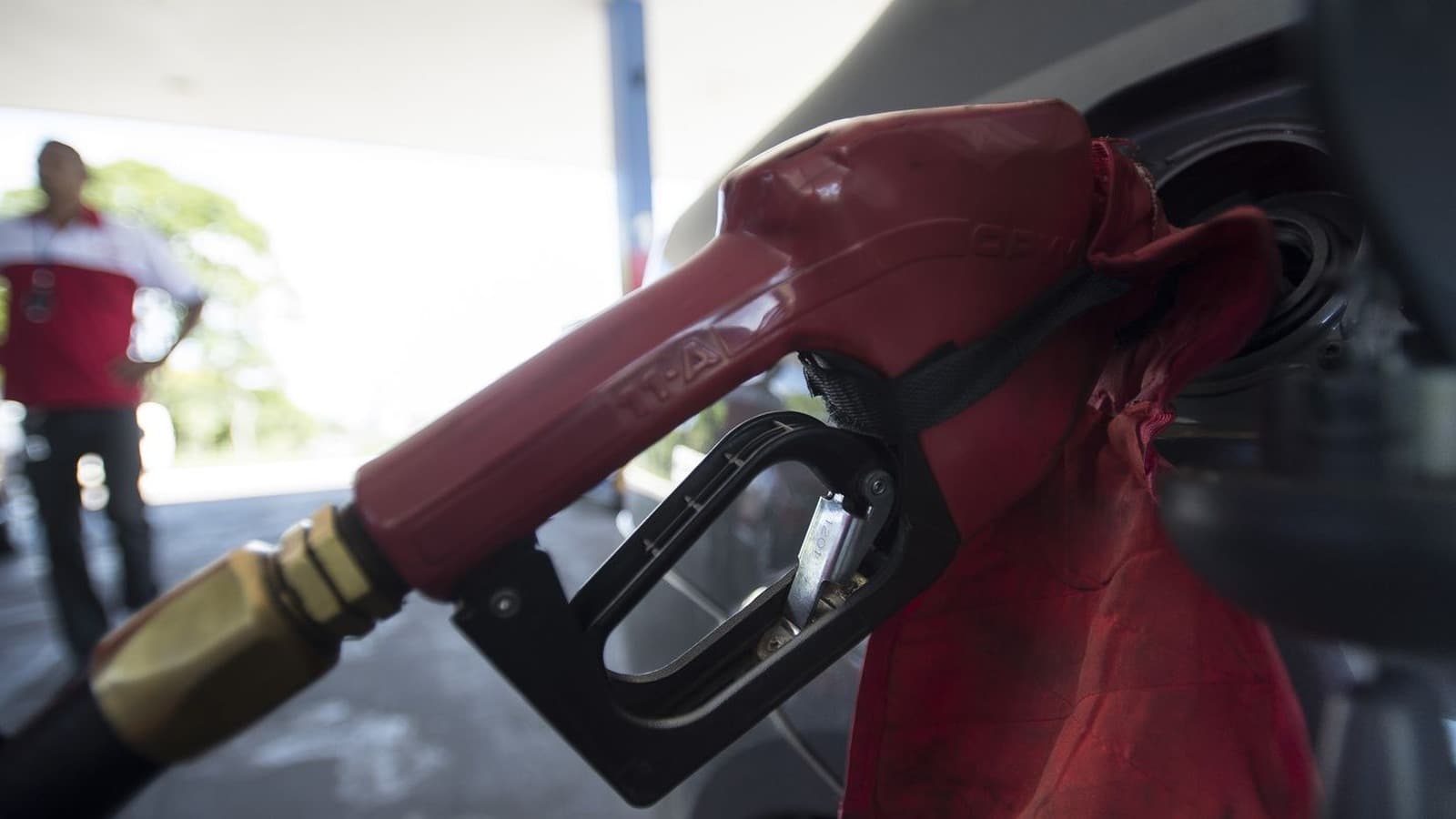 Foto de arquivo: bico de bomba de combustível abastecendo um veículo em um posto, com frentista de uniforme vermelho desfocado ao fundo