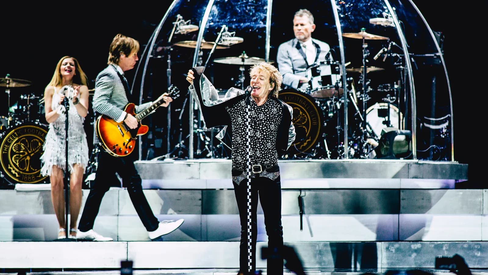 Rod Stewart canta em cima do palco durante show, com banda ao fundo
