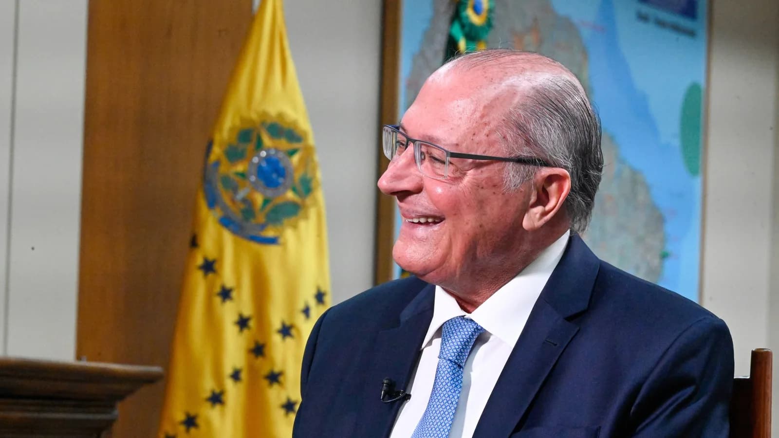 Geraldo Alckmin sorri durante entrevista, com bandeira do Brasil ao fundo