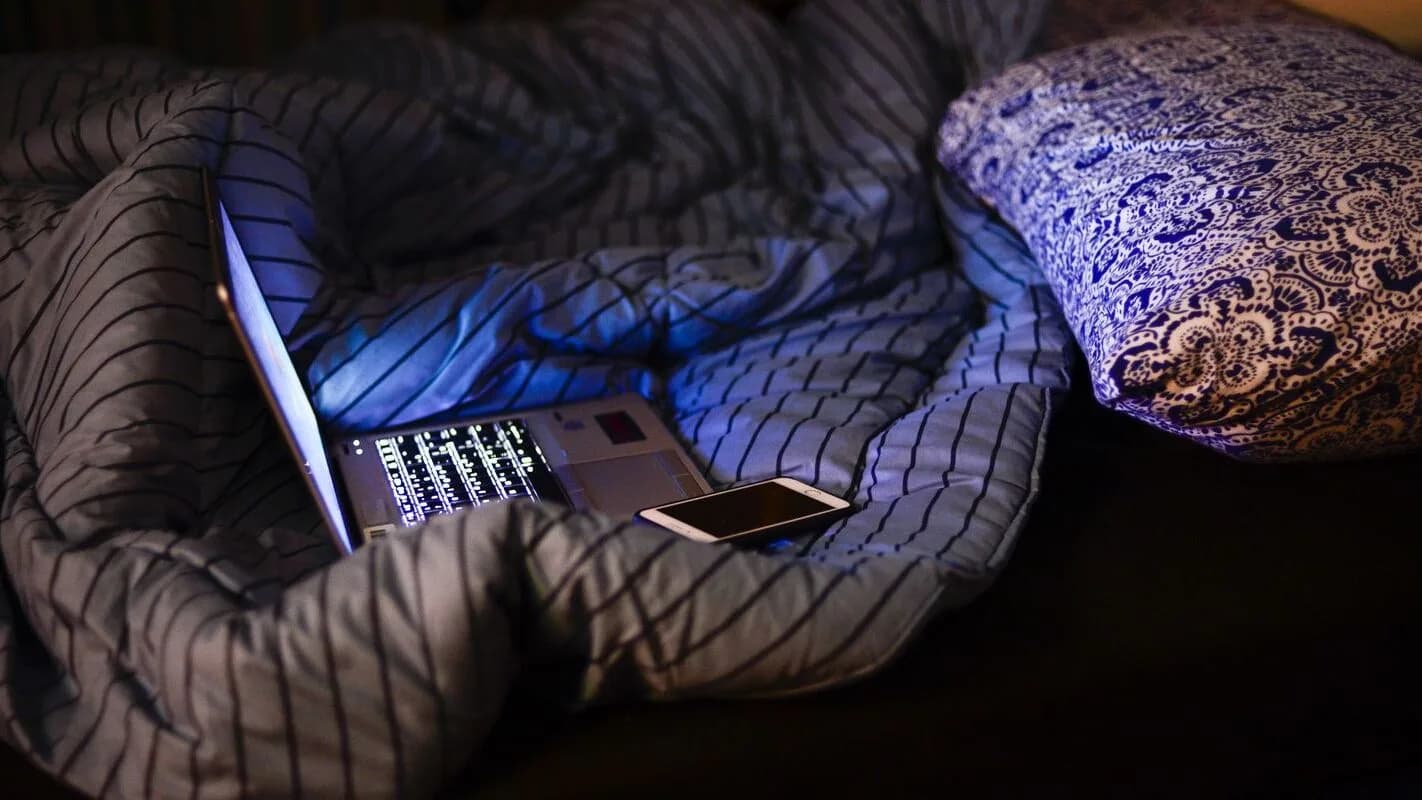 Pessoa dormindo sob os cobertores à noite, com celular e notebook ao lado da cama