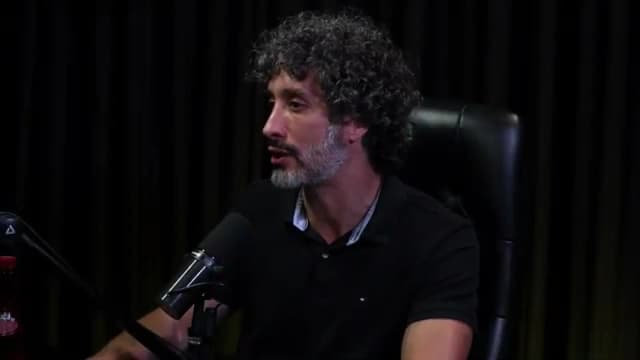 Carlos Eduardo Grimaldi fala recostado na cadeira, em perfil levemente lateral, em estúdio de podcast com cortina escura ao fundo