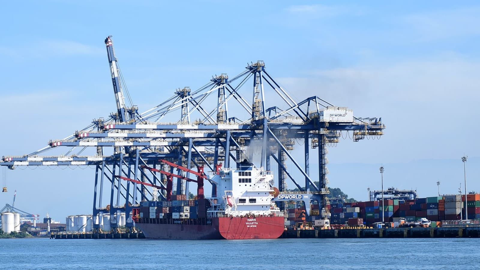 Navio cargueiro atracado sob guindastes de contêineres em terminal portuário, com pilhas de contêineres coloridos no cais; imagem de arquivo sobre comércio exterior