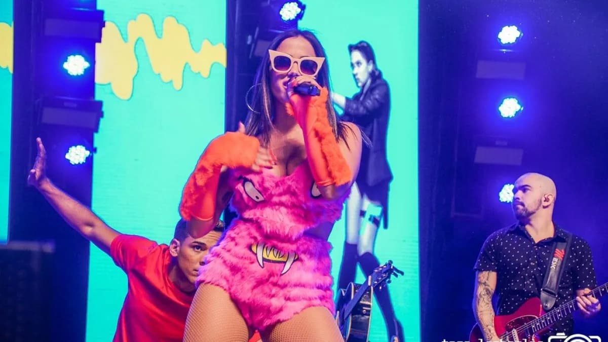 Anitta canta ao microfone durante show, usando fantasia rosa, em palco iluminado