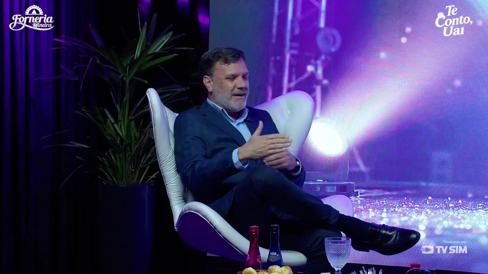 Alessandro Feijó, o Dr. LinkedIn, gesticula durante entrevista ao programa Te Conto, Uai, sentado em poltrona branca em estúdio com luzes roxas