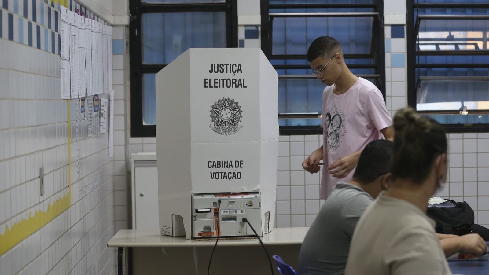 Cabina de votação da Justiça Eleitoral com urna eletrônica sobre a mesa, eleitor se aproximando e mesários sentados em uma seção eleitoral. Imagem de arquivo