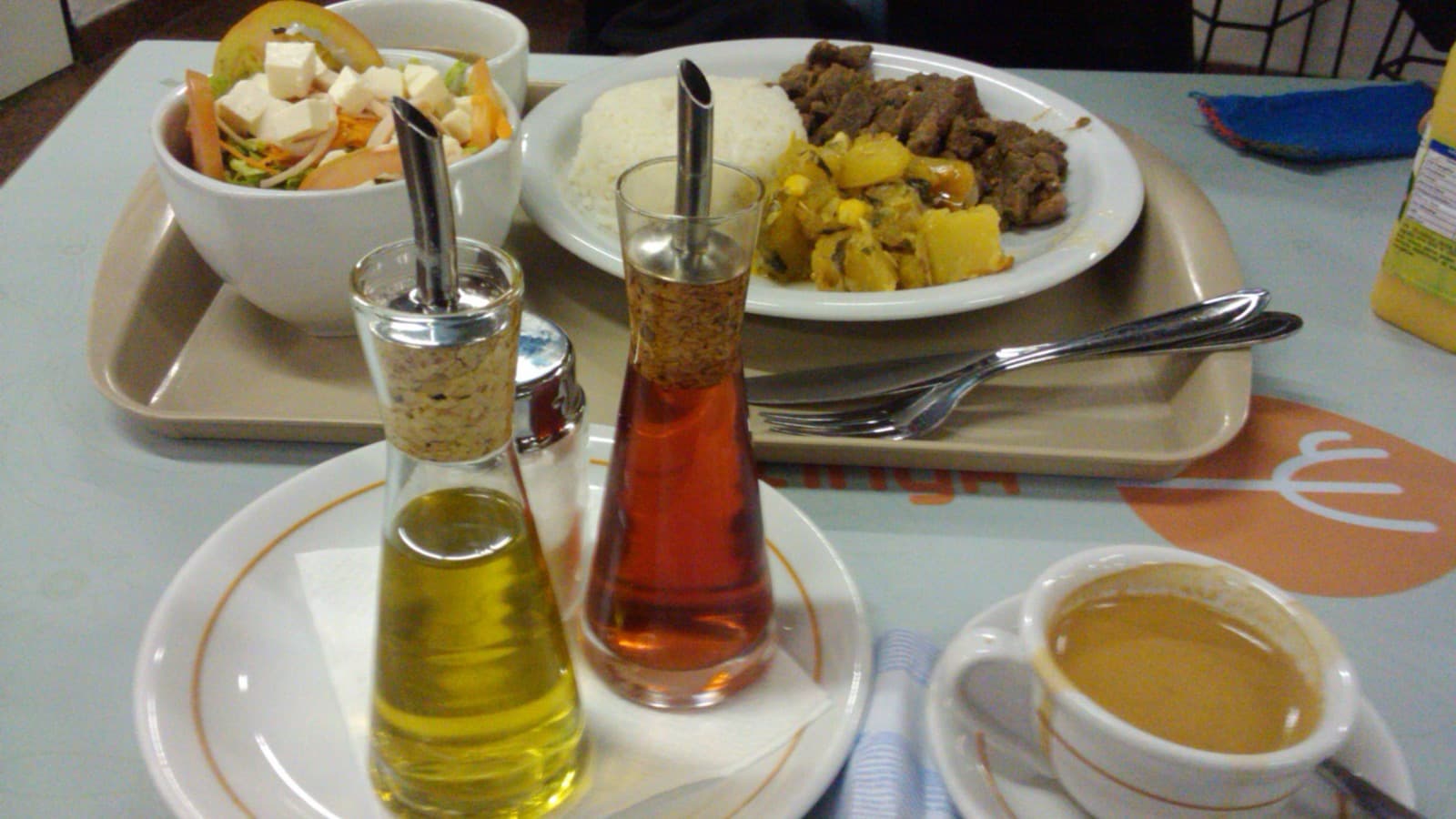 Bandeja de almoço em restaurante self-service com prato de arroz, carne e legumes, tigela de salada, galheteiro e xícara de café, em foto de arquivo