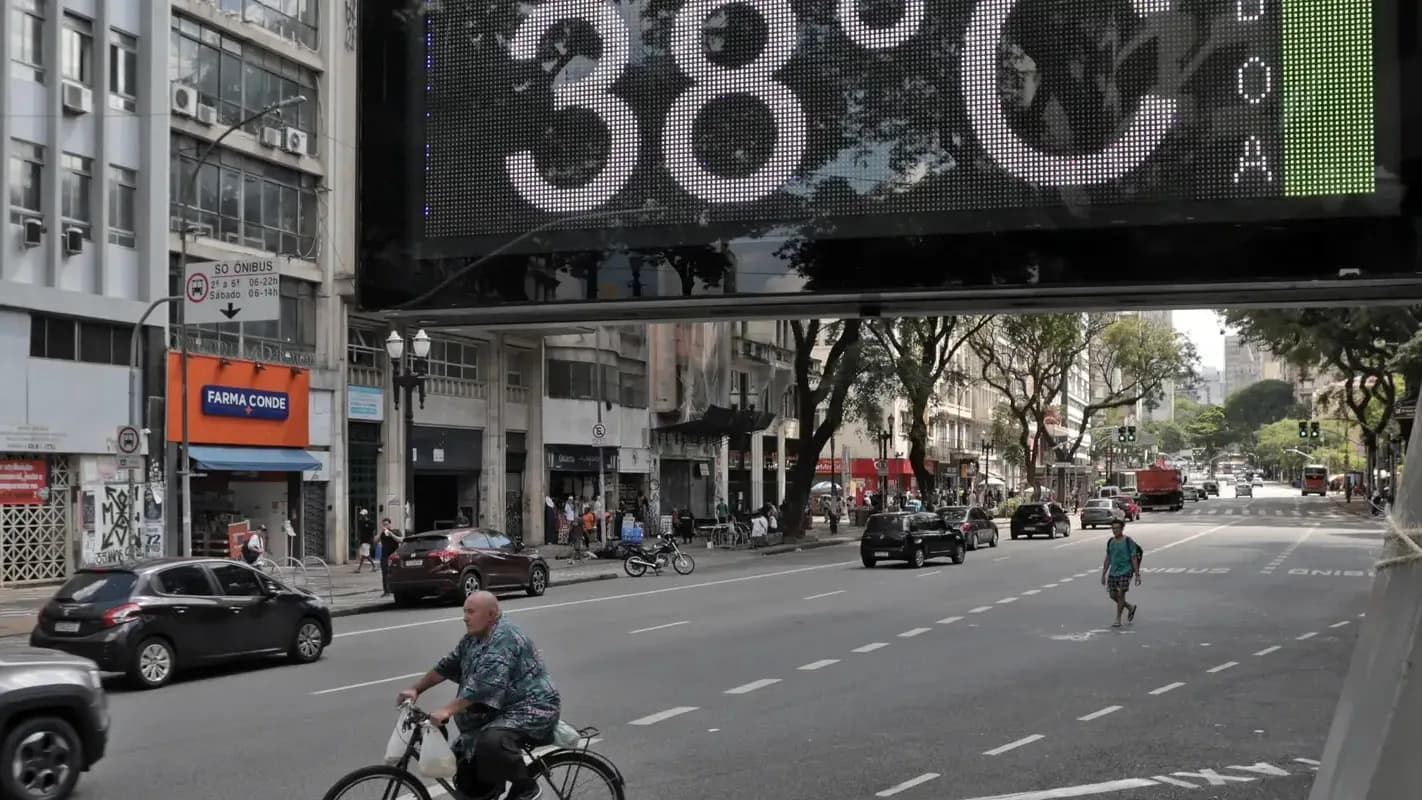 Termômetro de rua marca 38°C em avenida movimentada, com ciclista e pedestres passando