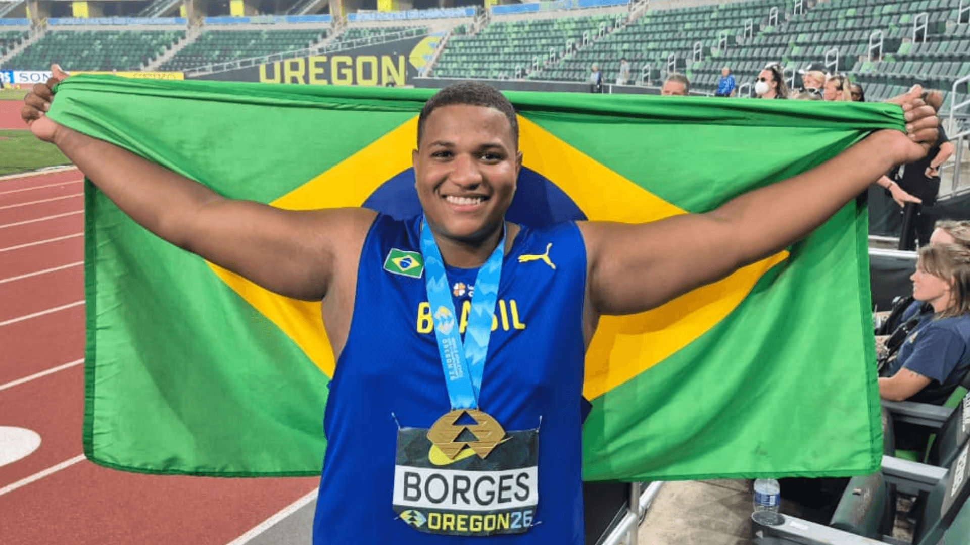 Alessandro Borges sorri segurando a bandeira do Brasil, com a medalha de ouro no peito, no estádio em Eugene