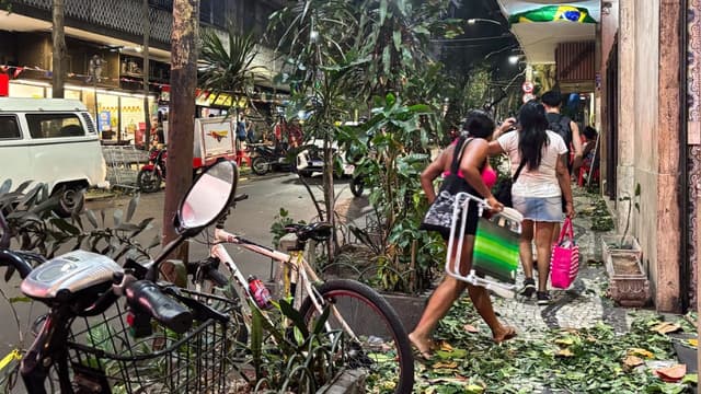 Pedestres caminham por calçada carioca coberta de folhas e galhos arrancados pela ventania, à noite, com bicicleta e moto encostadas e comércio ao fundo
