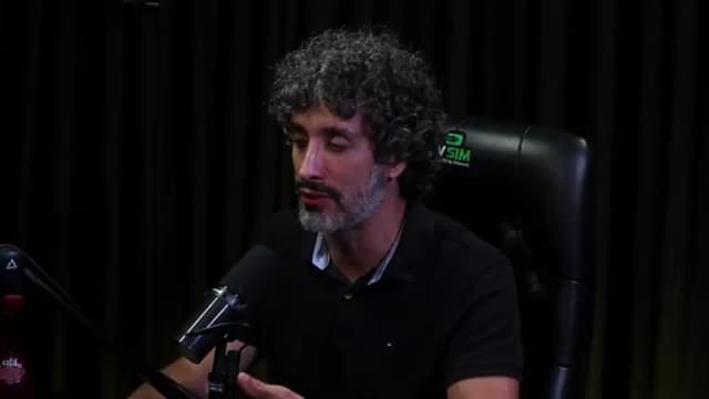 Carlos Eduardo Grimaldi fala olhando de lado ao microfone, com expressão séria, em estúdio de podcast com cortina escura ao fundo