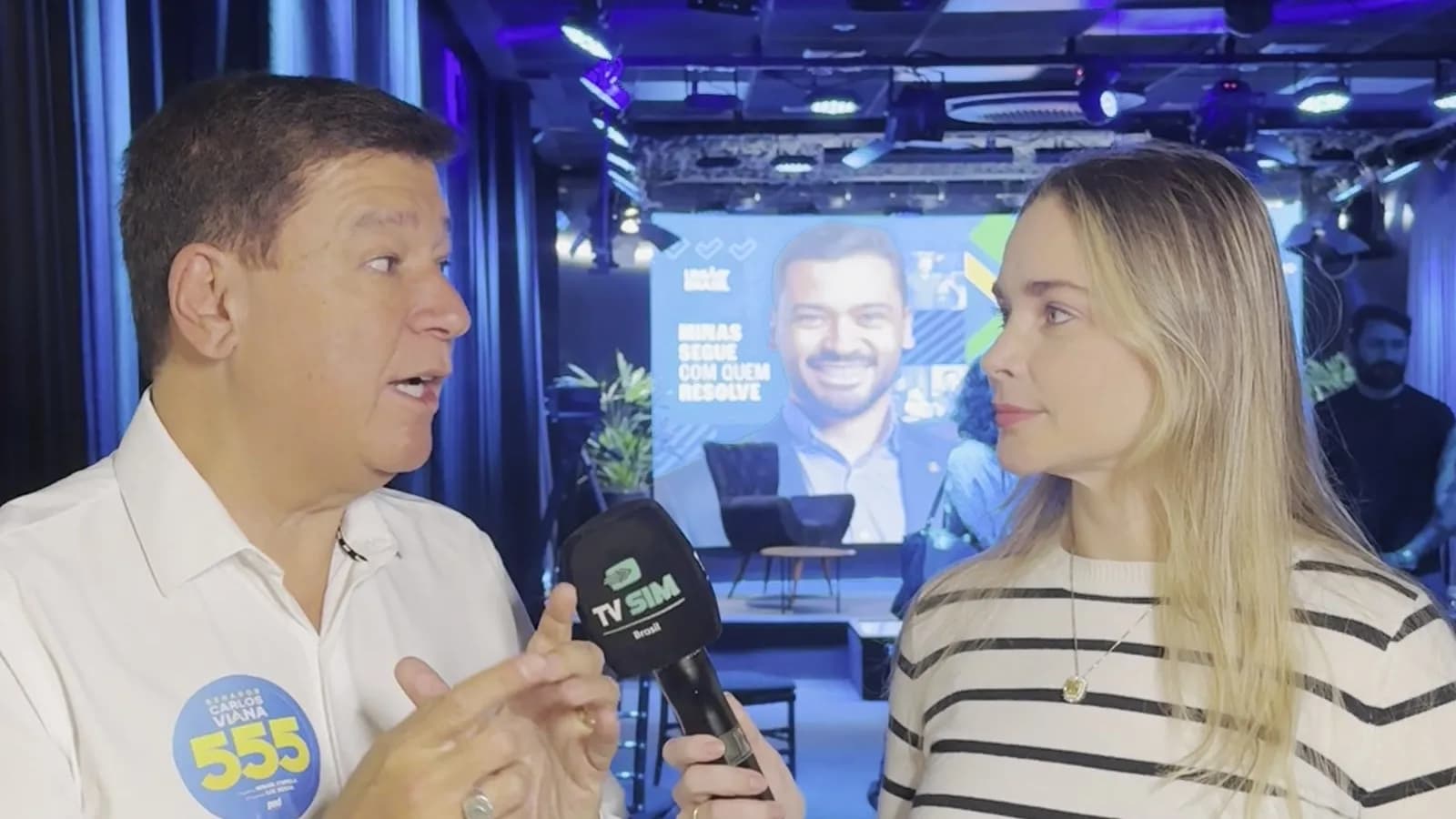 Carlos Viana gesticula ao falar ao microfone da TV Sim Brasil, ao lado da repórter, durante entrevista sobre sua candidatura ao Senado por Minas Gerais