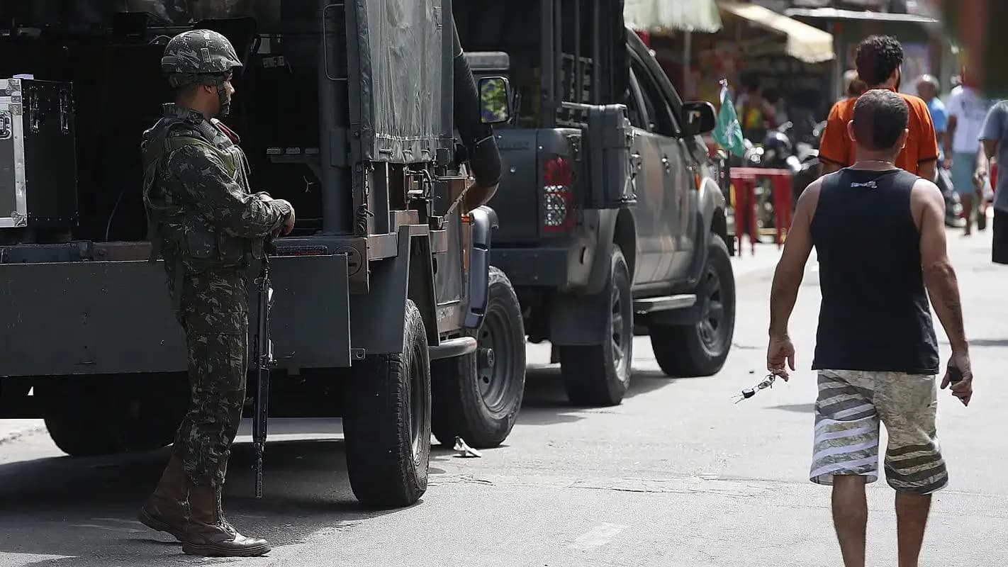 Militar armado ao lado de viatura blindada em rua do Rio de Janeiro, com moradores passando