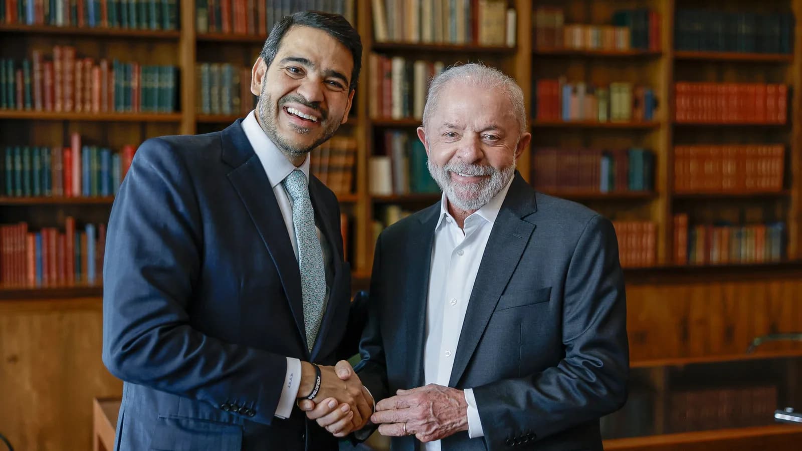 O presidente Lula cumprimenta Jorge Messias, sorridentes, em frente a uma estante de livros, no dia do anúncio da indicação ao STF