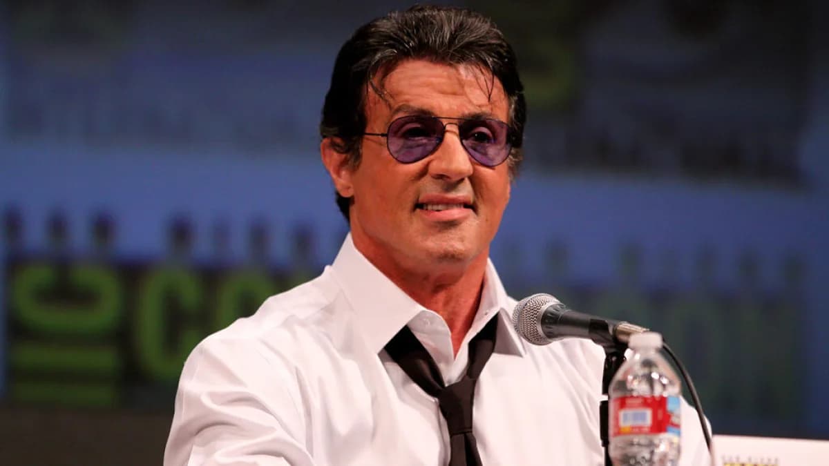 Sylvester Stallone usa óculos escuros durante evento público, sorrindo diante de microfone
