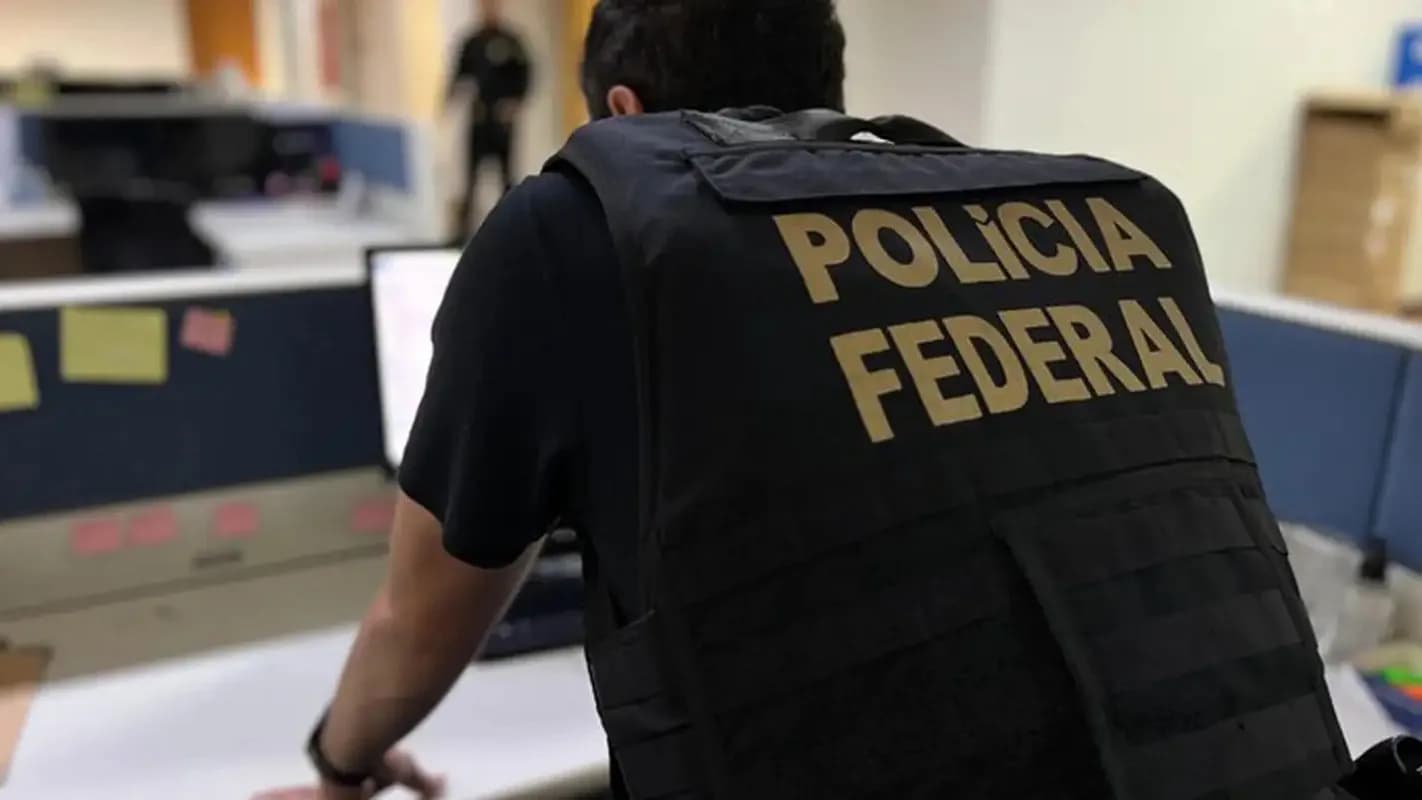 Agente da Polícia Federal com colete escrito Polícia Federal, visto de costas em escritório