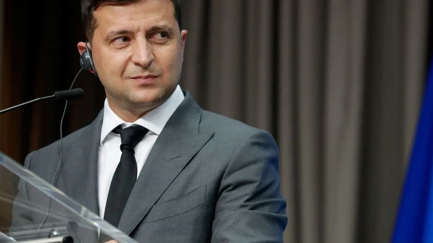 Volodymyr Zelensky fala ao microfone em pronunciamento, com bandeira da Ucrânia ao fundo