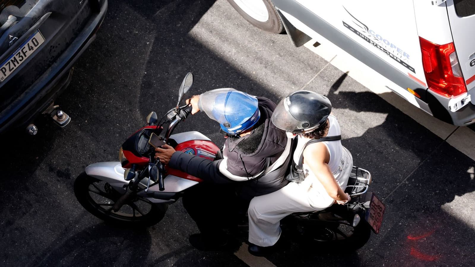 Vista de cima de uma motocicleta parada no trânsito entre carros, com piloto e passageiro usando capacete aberto; imagem de arquivo