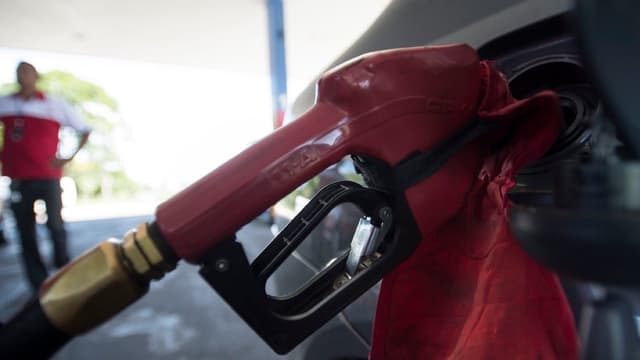 Bico de combustível vermelho abastecendo um veículo em um posto, com um frentista de uniforme desfocado ao fundo, em foto de arquivo