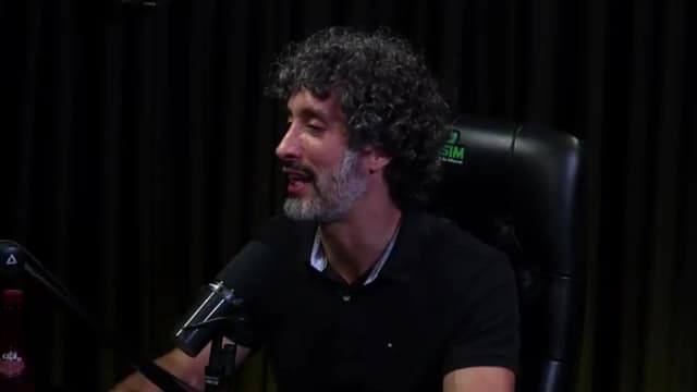 Carlos Eduardo Grimaldi sorri enquanto fala ao microfone, olhar erguido, em estúdio de podcast com cortina escura ao fundo