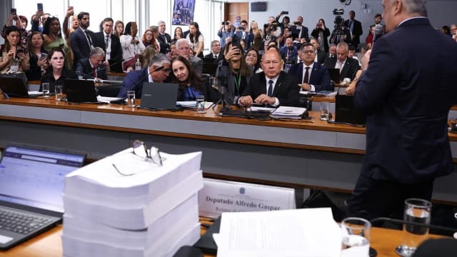 Sessão da CPMI do INSS lotada, com pilha de documentos do relatório sobre a mesa