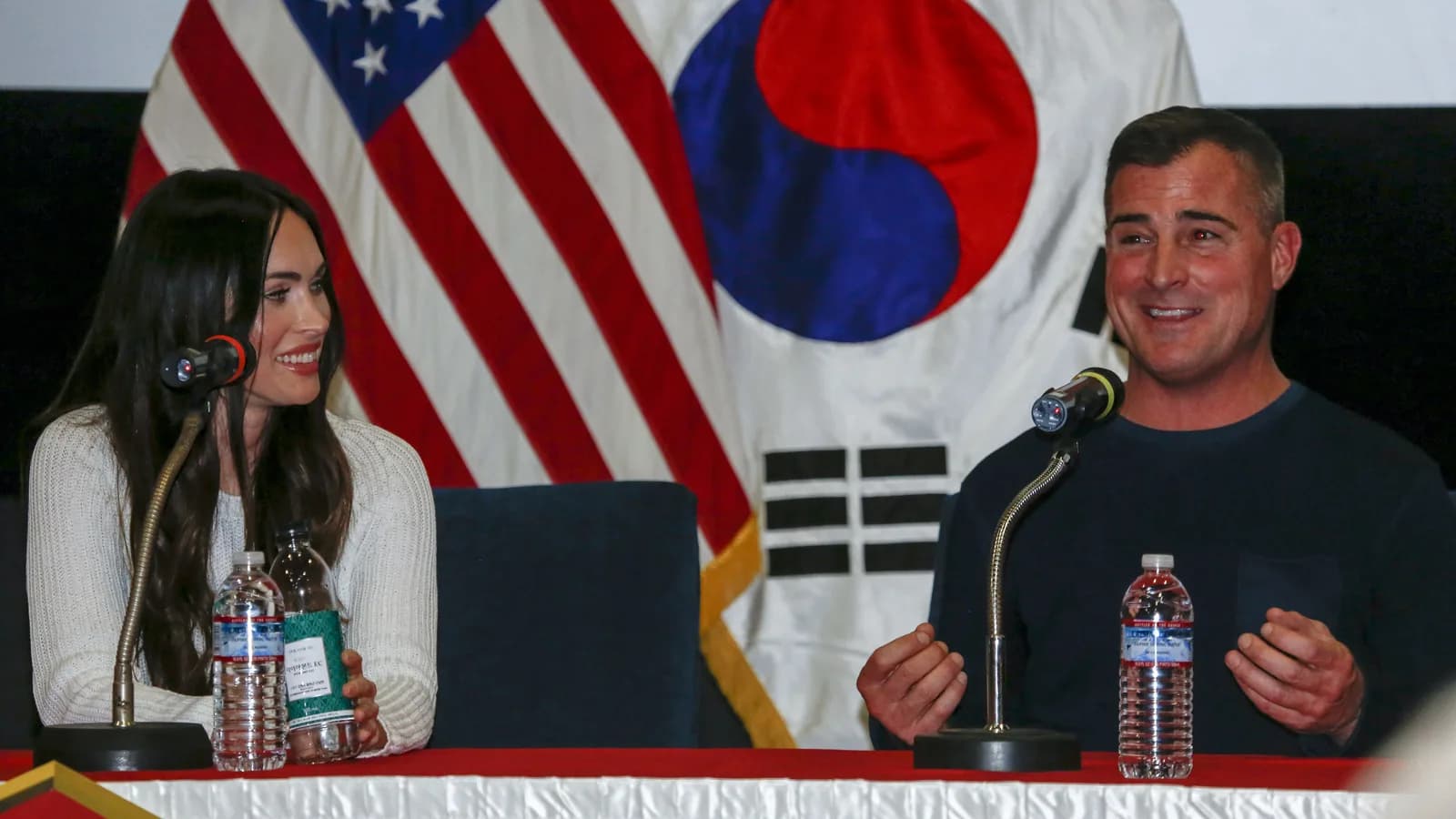 Megan Fox sorri em painel público, sentada à mesa com microfone
