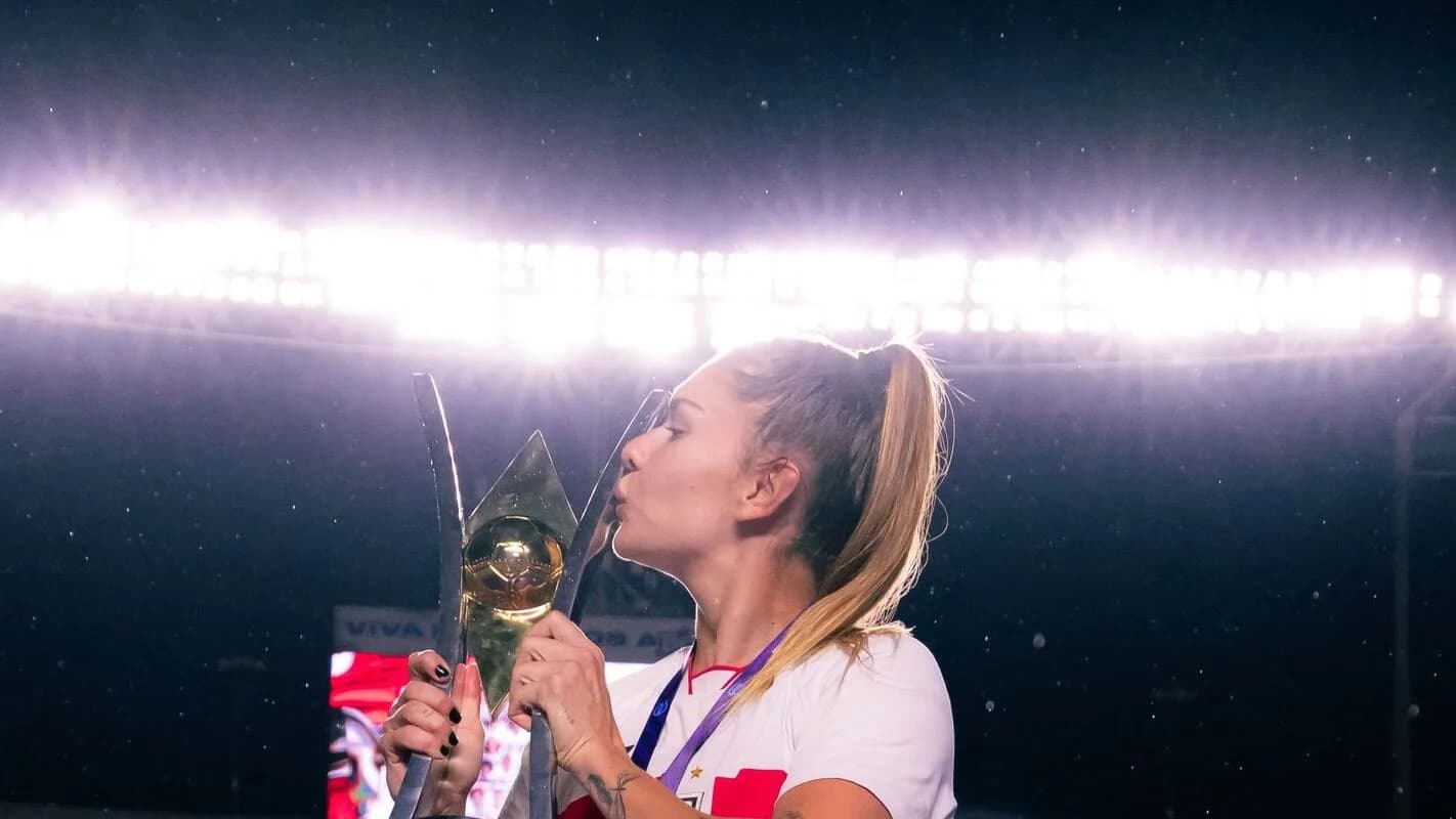 A jogadora Carol Gil, de costas para o gramado iluminado do estádio, beija o troféu de campeã
