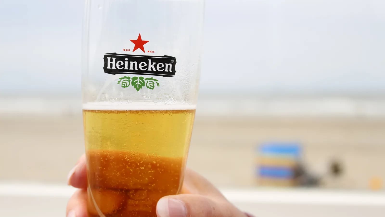 Mão segurando um copo de cerveja Heineken com praia ao fundo