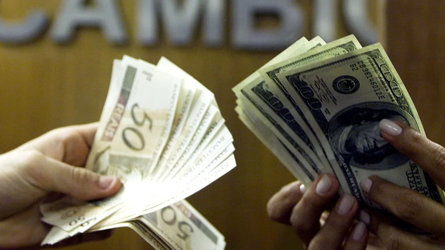 Mãos trocam notas de real e dólar, ilustrando operação financeira