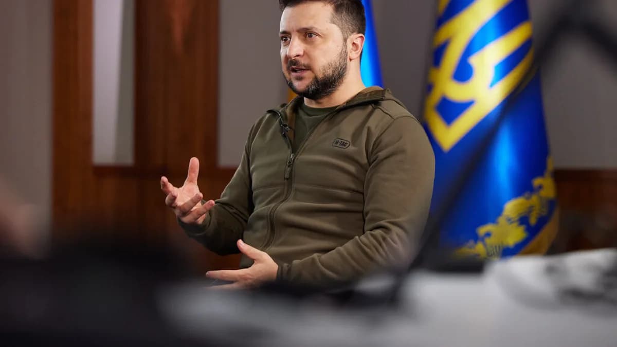 Volodymyr Zelensky gesticula durante entrevista, vestindo moletom verde-militar, com bandeiras da Ucrânia ao fundo