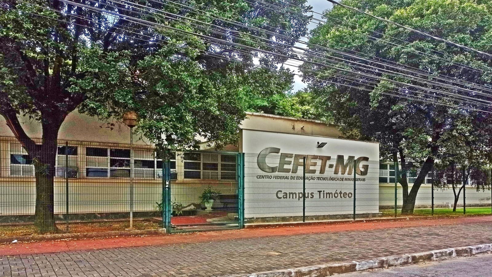 Fachada do campus Timóteo do Cefet-MG, com letreiro em relevo escrito CEFET-MG, Centro Federal de Educação Tecnológica de Minas Gerais, Campus Timóteo, atrás de um gradil verde e de árvores na calçada