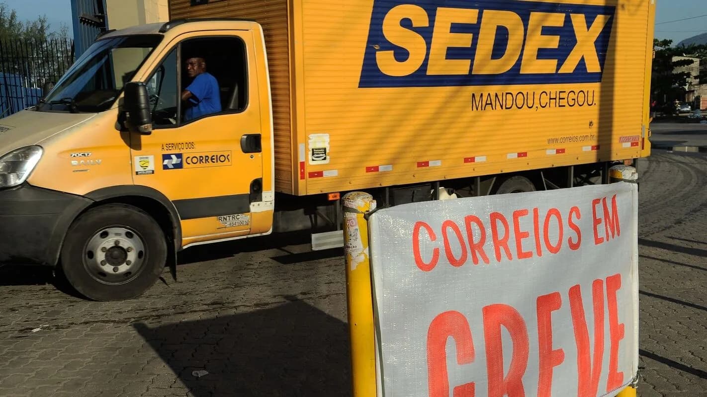 Caminhão Sedex dos Correios ao lado de faixa com a mensagem "Correios em greve", em ato de trabalhadores contra mudanças no plano de saúde
