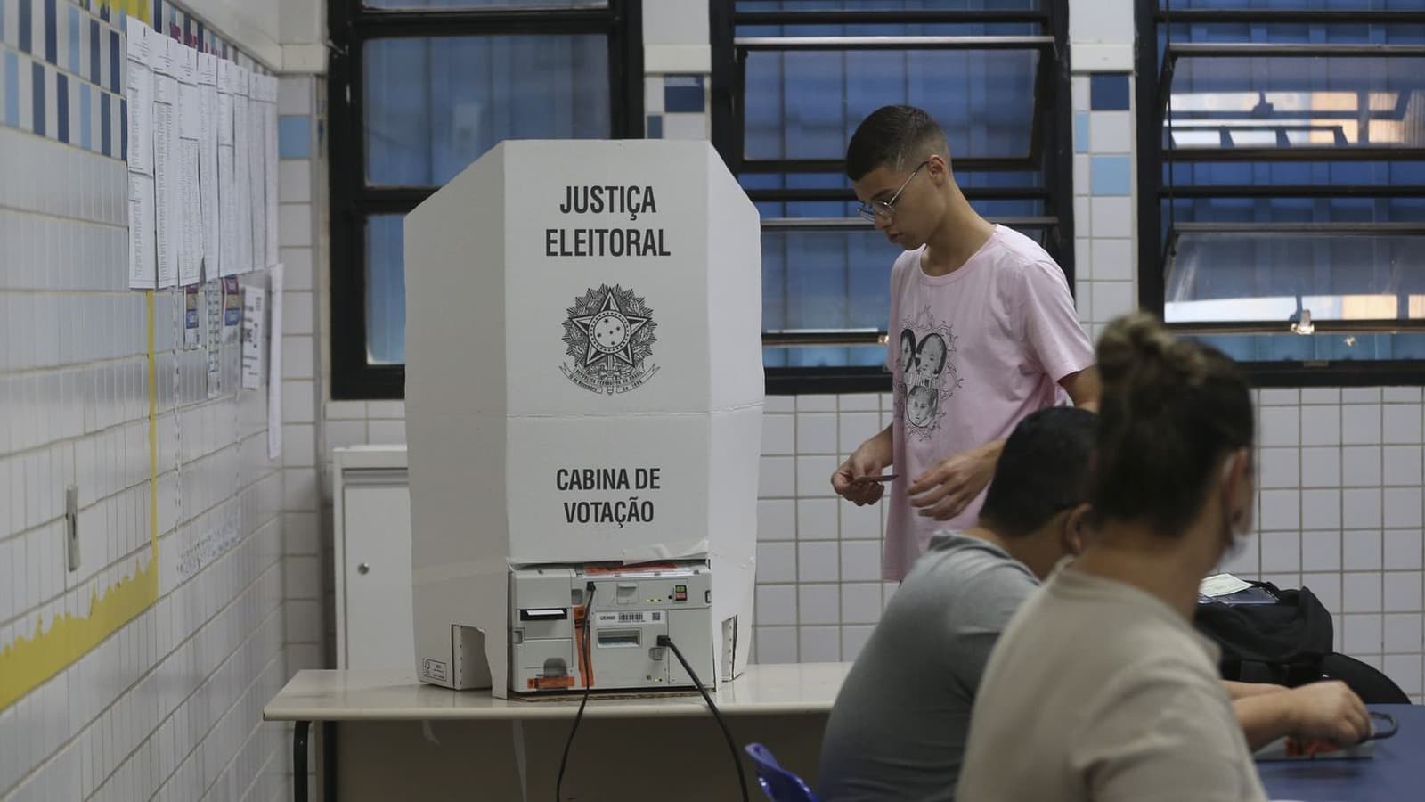 Eleitor se aproxima de uma cabina de votação da Justiça Eleitoral em uma seção eleitoral, com mesários à mesa; imagem de arquivo de pleito anterior