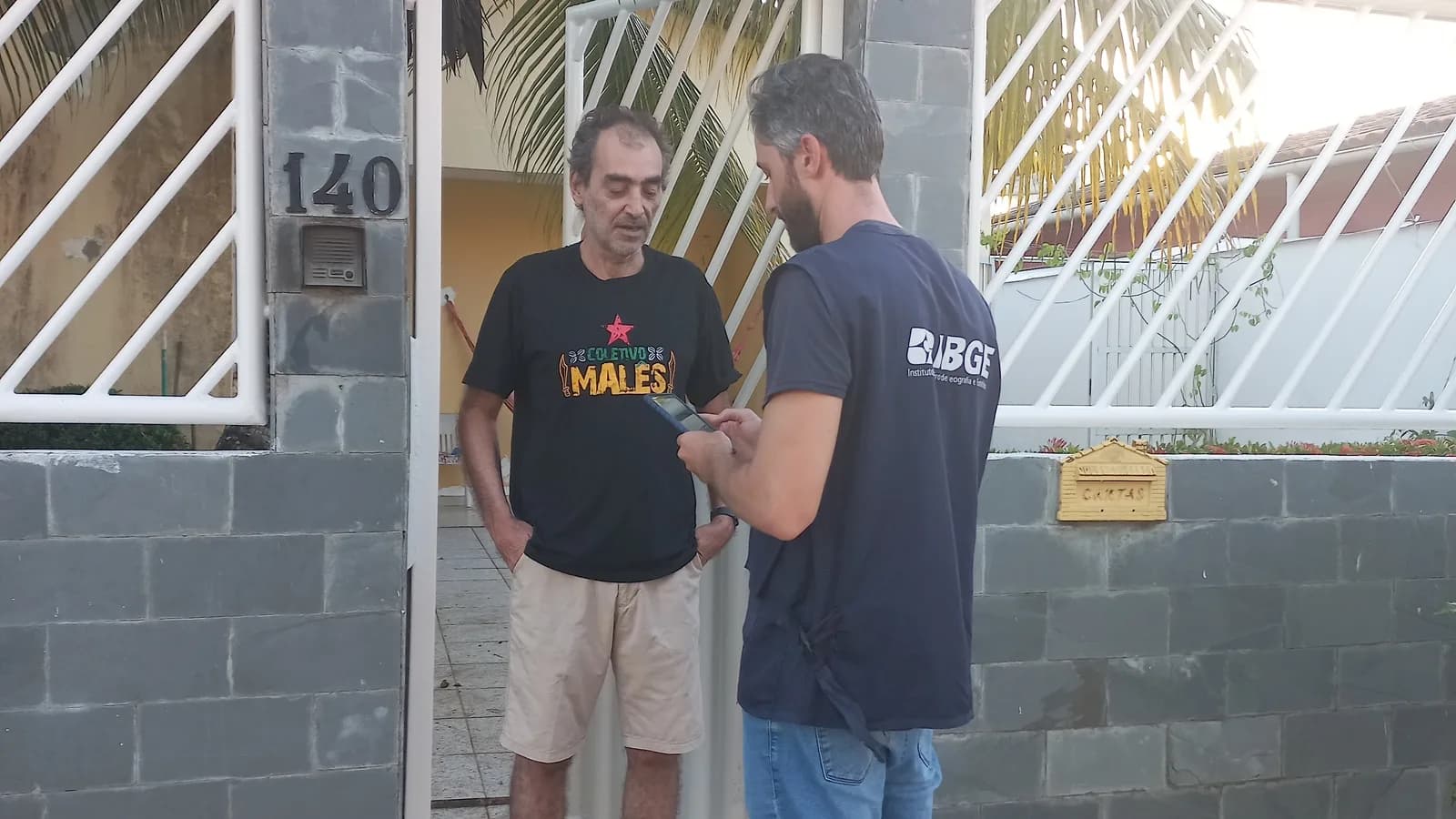 Recenseador do IBGE, de uniforme, entrevista morador na porta de casa