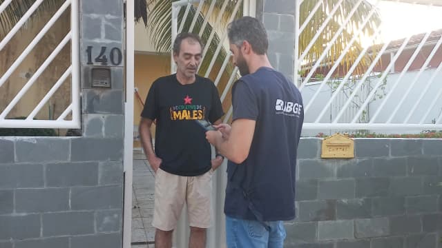 Recenseador do IBGE, de uniforme, entrevista morador na porta de casa
