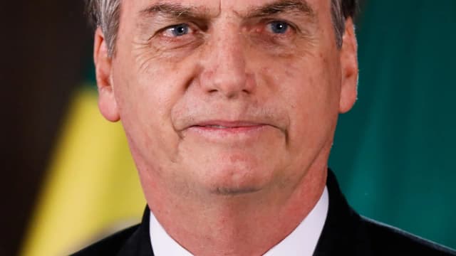 Jair Bolsonaro em retrato oficial, de terno escuro, com bandeiras do Brasil ao fundo