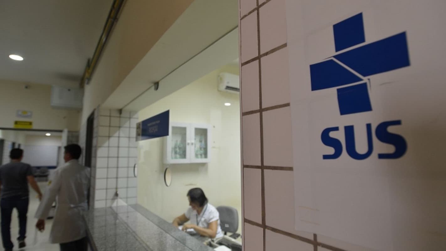 Logo do SUS na parede de corredor de unidade de saúde, com profissional de jaleco e atendente ao fundo (foto de arquivo)