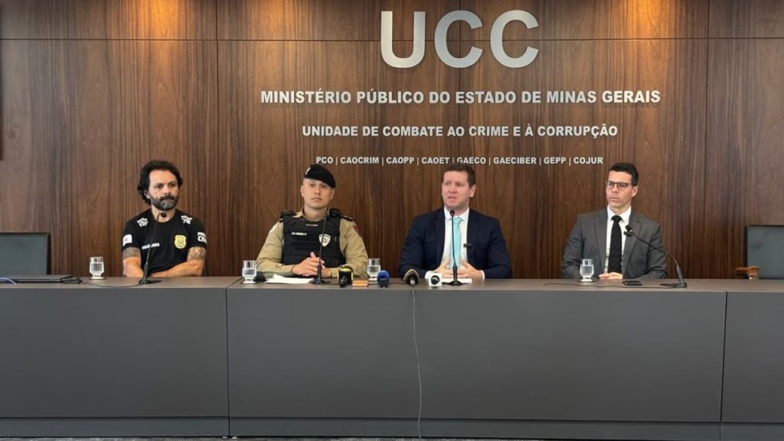 Delegado, capitão da PM e dois promotores do MPMG sentados em coletiva de imprensa, com painel da Unidade de Combate ao Crime e à Corrupção ao fundo