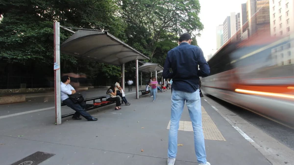Pessoas aguardam em ponto de ônibus enquanto veículo se aproxima, em rua de cidade brasileira