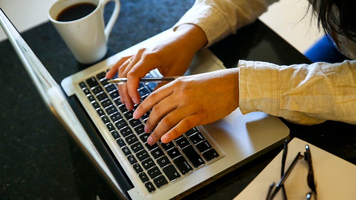 Mãos de uma pessoa digitando em um notebook sobre uma bancada, ao lado de uma xícara de café e de um par de óculos