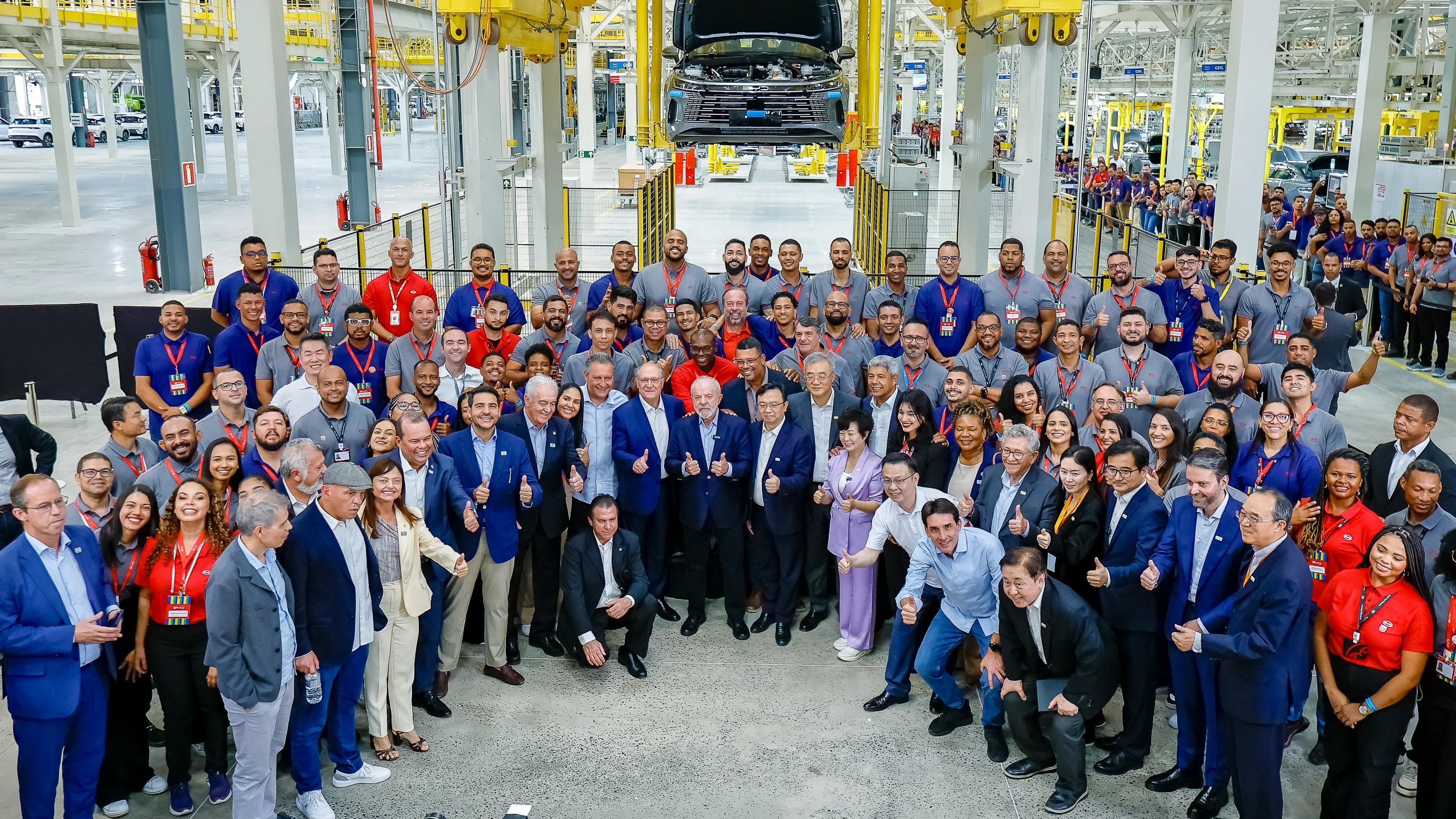 O presidente Lula, cercado por dezenas de trabalhadores brasileiros e executivos chineses, faz sinal de positivo na linha de montagem da fábrica da BYD em Camaçari, com um veículo suspenso ao fundo