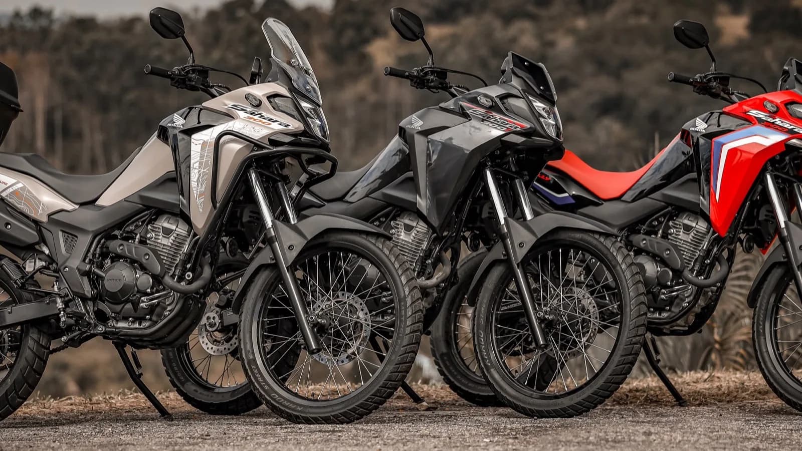 Três motocicletas Honda Sahara 300 2027 enfileiradas ao ar livre, em tons de cinza claro, cinza-escuro e vermelho