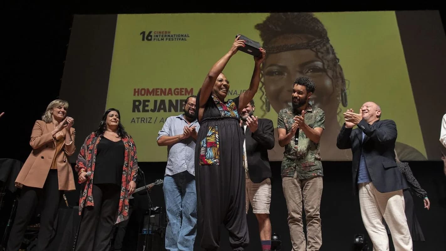 Atriz Rejane Faria recebe homenagem em pé no palco, aplaudida por outras pessoas, com seu retrato projetado ao fundo