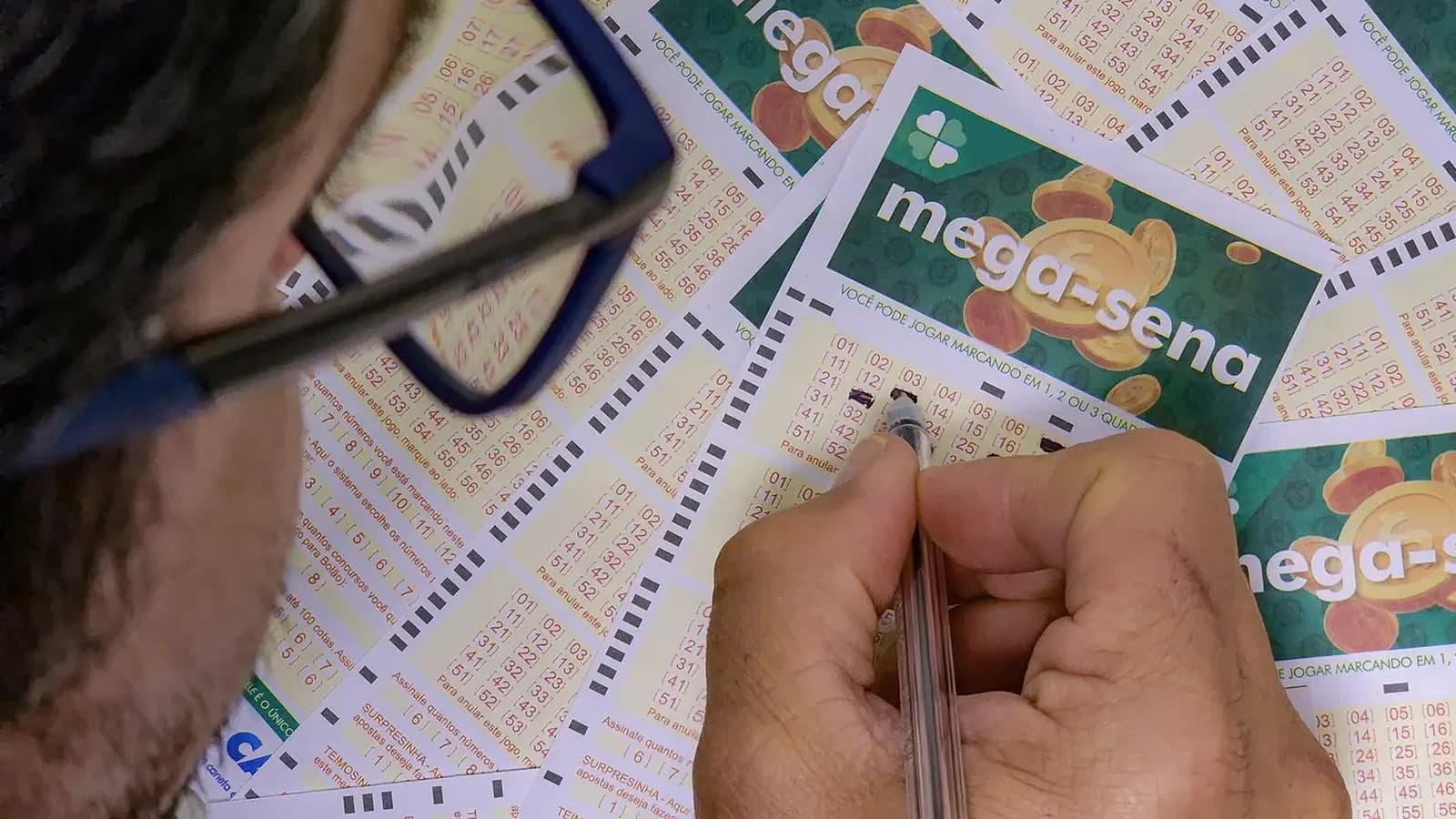 Apostador de óculos preenche com caneta volantes da Mega-Sena espalhados sobre uma mesa; imagem de arquivo
