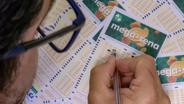 Apostador de óculos preenche com caneta volantes da Mega-Sena espalhados sobre uma mesa; imagem de arquivo