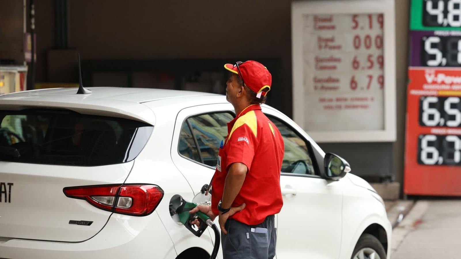 Frentista de uniforme vermelho e boné abastece um carro branco em posto de combustível, com o painel de preços ao fundo. Imagem de arquivo