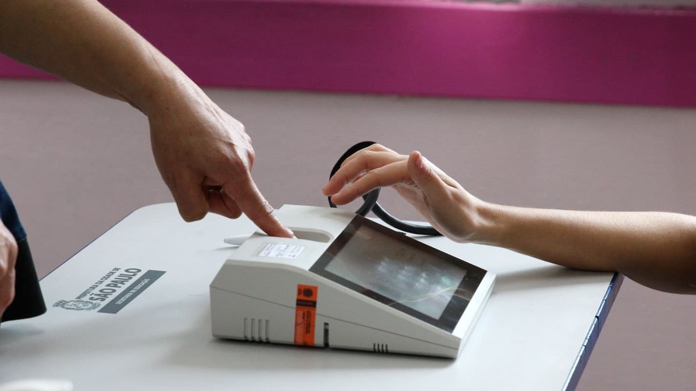 Duas mãos sobre um leitor biométrico apoiado em mesa de seção eleitoral, uma delas encostando o dedo no equipamento; imagem de arquivo