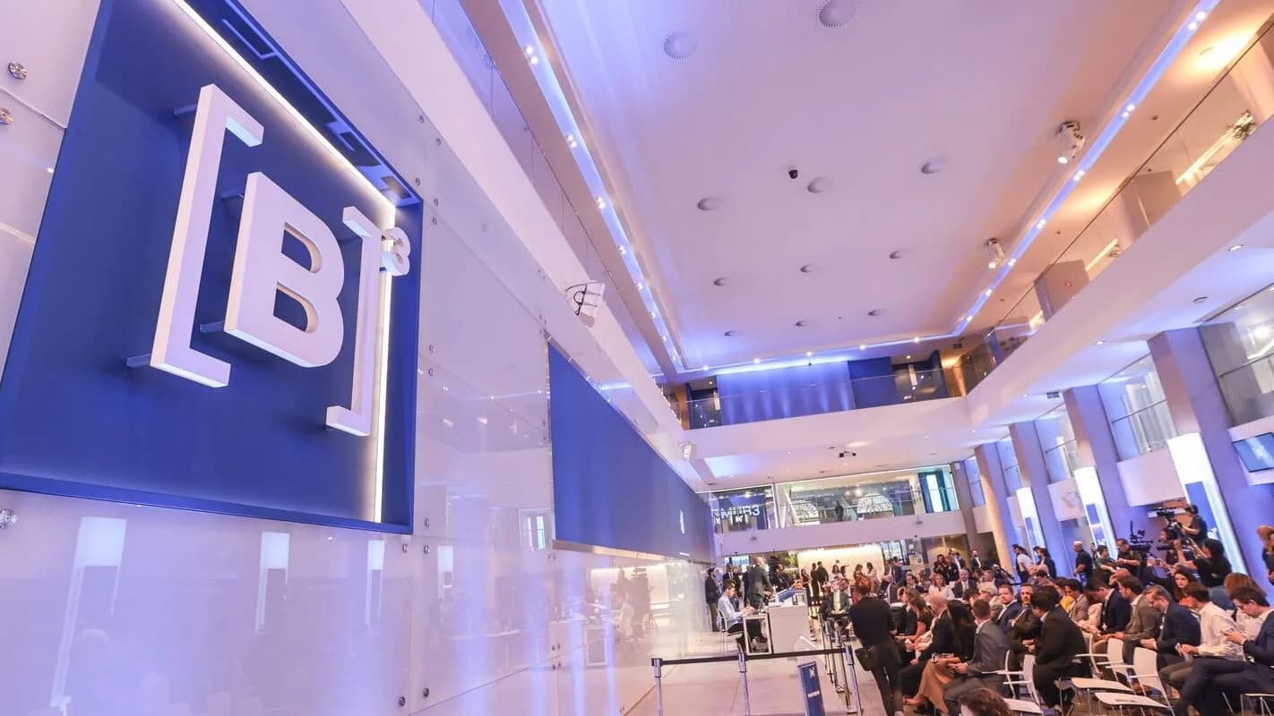 Painel com o logo da B3 na sede da bolsa de valores em São Paulo, com público sentado ao fundo