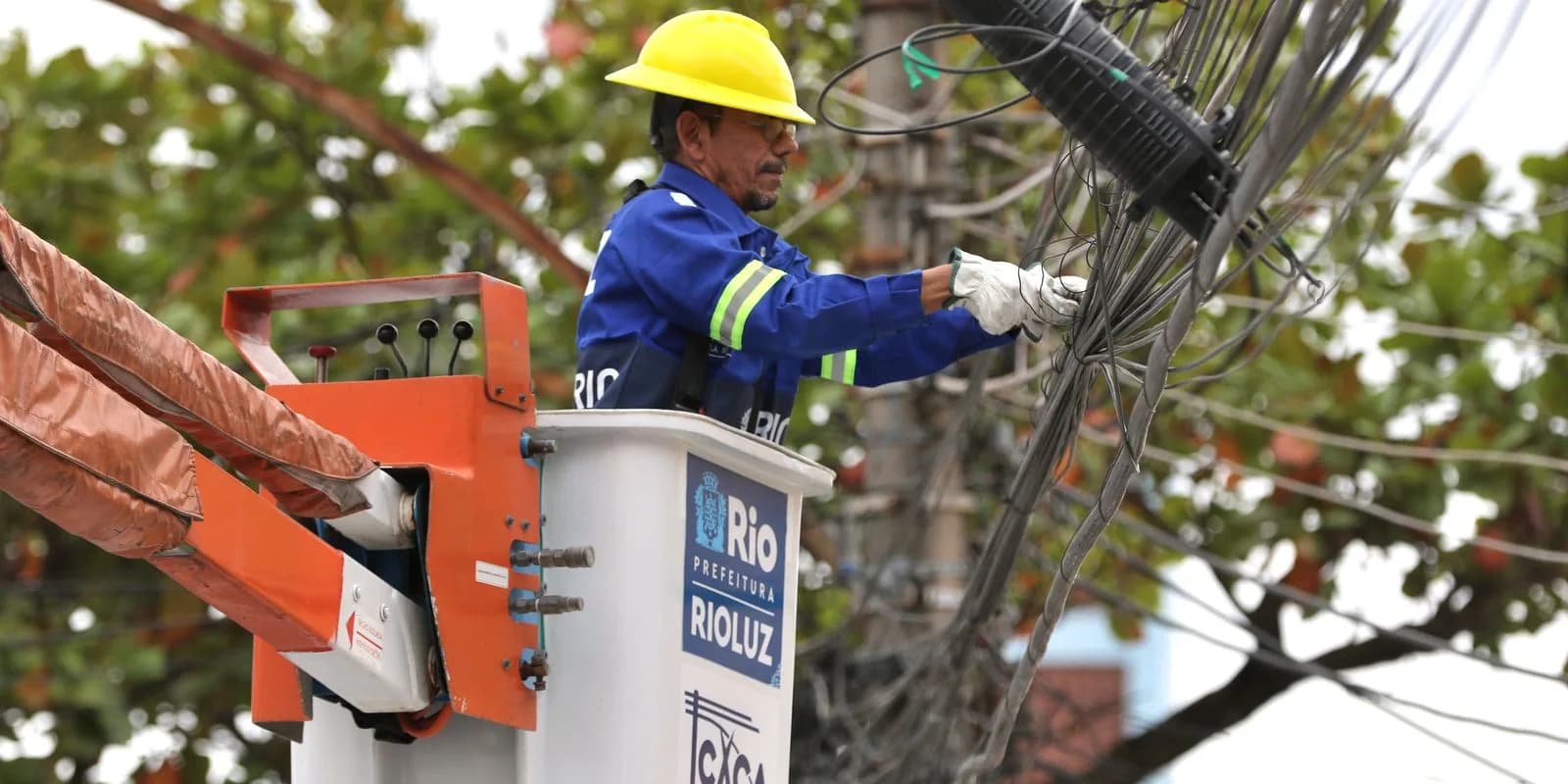 Eletricista trabalha em rede de fiação elétrica sobre poste, com capacete e luvas de proteção (foto de arquivo)